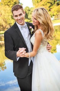 Married couple on their wedding day holding hands next to pond while bride looks lovingly at groom and groom smiles at camera
